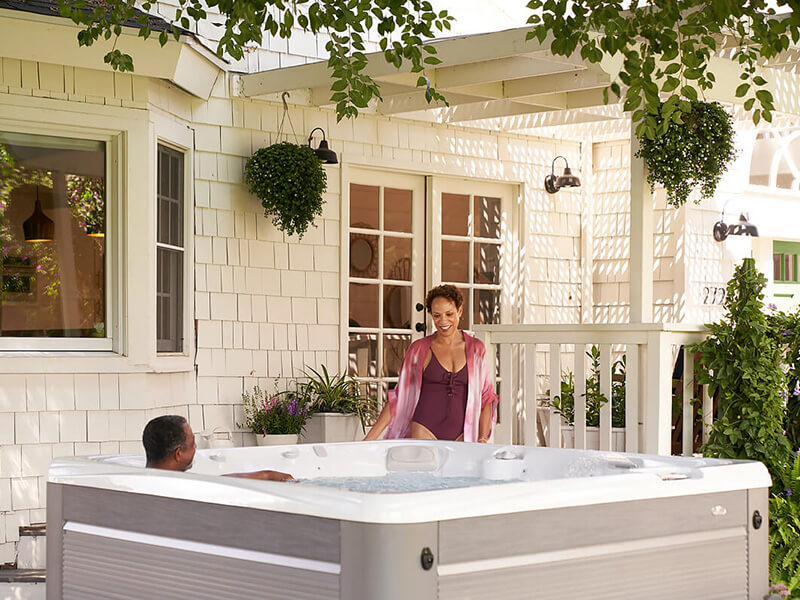 A couple enjoying a Caldera Spas hot tub in a cozy backyard setting. The man is relaxing in the warm, bubbling water, while the woman, wearing a swimsuit and a light cover-up, is smiling as she prepares to join him.